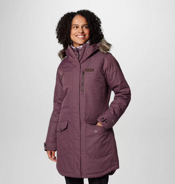 A model in a mulberry-colored winter parka with a fur-lined hood, flashing a smile. The coat boasts a sleek, modern silhouette with functional front pockets.