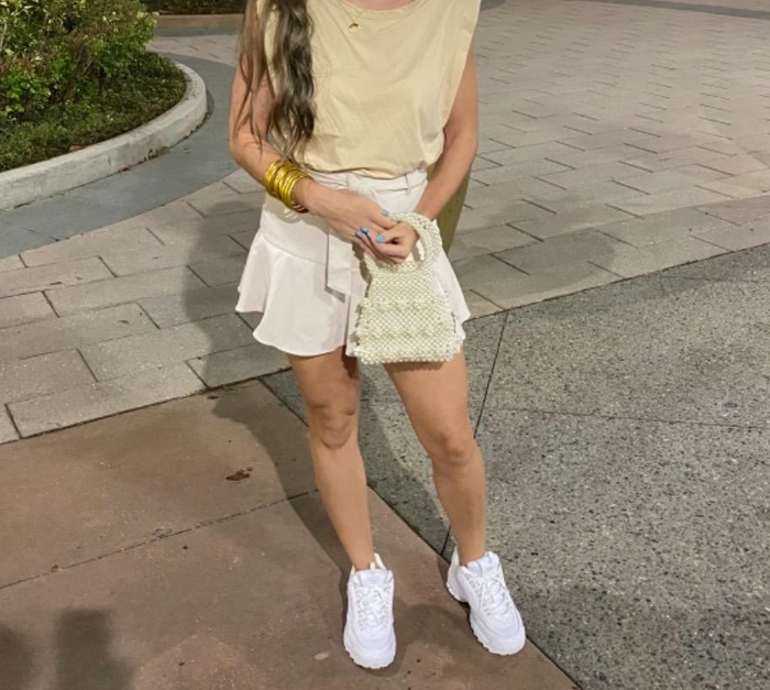 A reviewer wearing white sneakers while holding a small woven handbag
