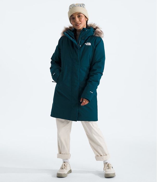 Person wearing a teal fur-trimmed parka, knit hat, and casual pants, showing off winter fashion