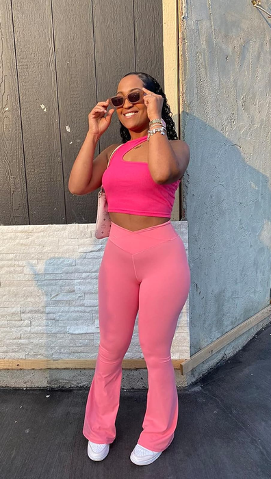 A fashionably dressed individual in a pink top and flared pants, wearing sunglasses, posing outdoors near a building