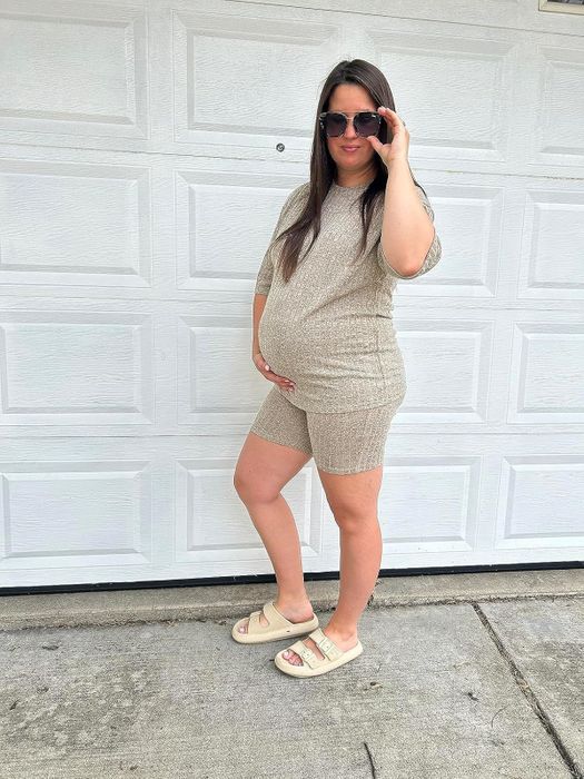 A pregnant reviewer in matching loungewear and sunglasses stands in front of a garage door, wearing comfortable sandals. The image captures a relaxed, at-home shopping vibe