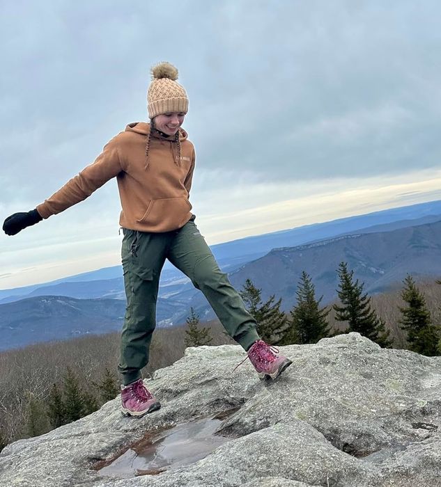 A reviewer wearing hiking pants and a beanie, balancing on a mountain rock with a breathtaking view in the background