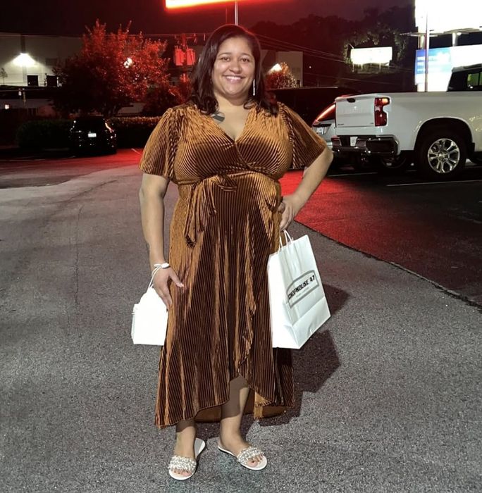 A woman smiles brightly while holding shopping bags in a nighttime parking lot. She’s dressed in a pleated dress and sandals, with a glowing red neon sign in the background