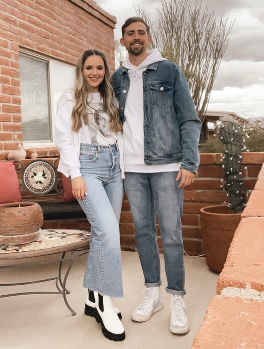 Two individuals dressed in casual denim outfits, including jeans and jackets, standing near a rustic outdoor seating area