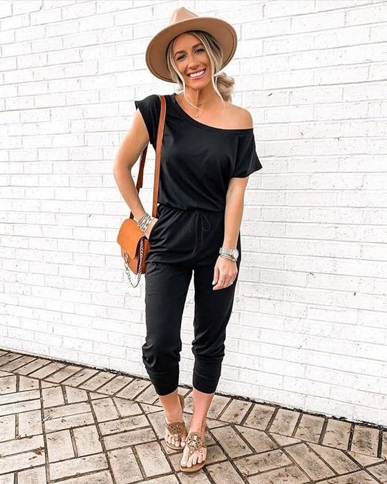 A model styled in the black jumpsuit, accessorized with a wide-brimmed hat, sandals, and a crossbody bag