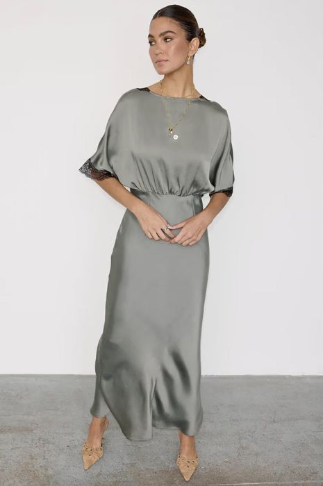 A person models a sophisticated satin dress adorned with lace trim, accessorized with statement earrings and a pendant necklace, gazing to the side