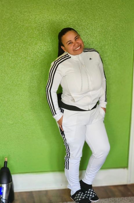 A reviewer in a white Adidas tracksuit with stripes, leaning against a green wall and smiling