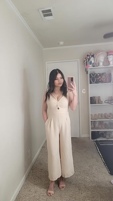 A reviewer takes a mirror selfie wearing a sleeveless beige jumpsuit and heels, standing in a room with a shelf showcasing handbags and shoes