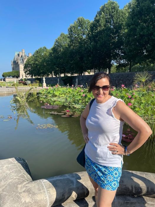 A reviewer wearing a sleeveless white top and a blue patterned skirt stands by a pond with lily pads and a stone embankment, smiling at the camera