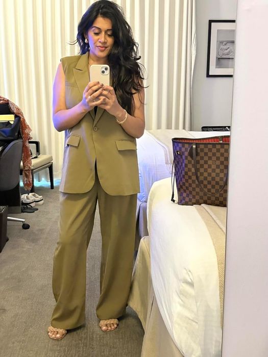 A reviewer takes a mirror selfie in a hotel room, wearing a loose-fitting sleeveless suit and holding a smartphone. A designer handbag is visible on the bed