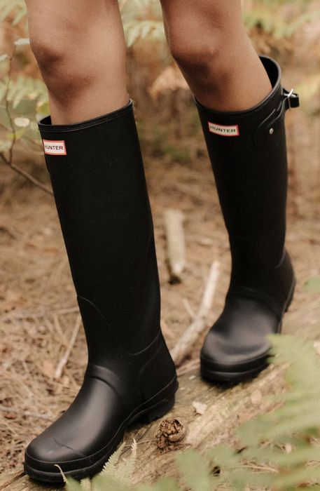 A person standing in a forest wearing tall black rubber boots, surrounded by lush foliage and branches
