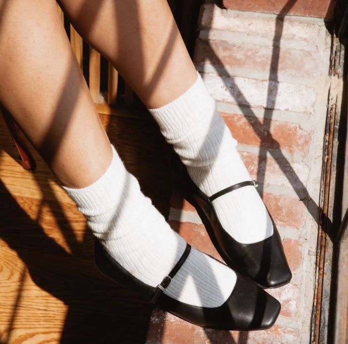 A person wearing white ribbed socks and black Mary Jane flats, seated on a wooden floor with sunlight creating soft shadows