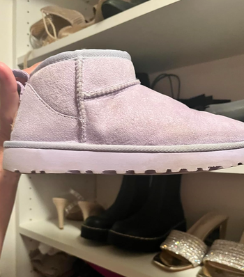 A hand holding a single suede slip-on boot in front of a shoe closet filled with various footwear styles, including high heels