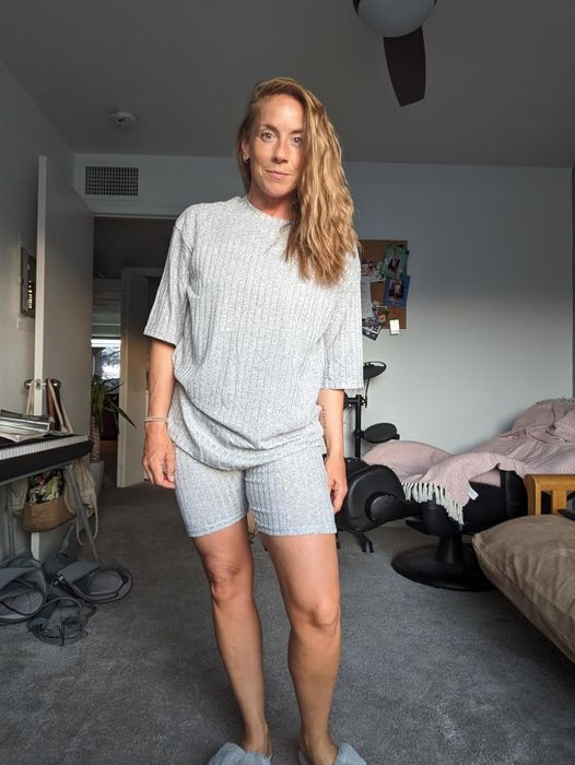 reviewer in cozy gray loungewear stands in a room featuring a bed, chair, and keyboard