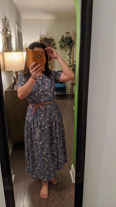 A reviewer takes a mirror selfie wearing a floral-patterned, calf-length dress belted at the waist, standing in a home setting. Their face is obscured by the phone
