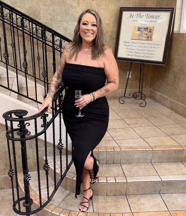 An individual in a chic black strapless dress holding a glass, standing on a staircase with a sign in the background displaying venue and rental information