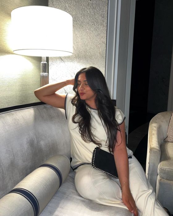 A reviewer in a chic white dress with black trim sits on a couch beside a lit lamp, posing with her hand behind her head