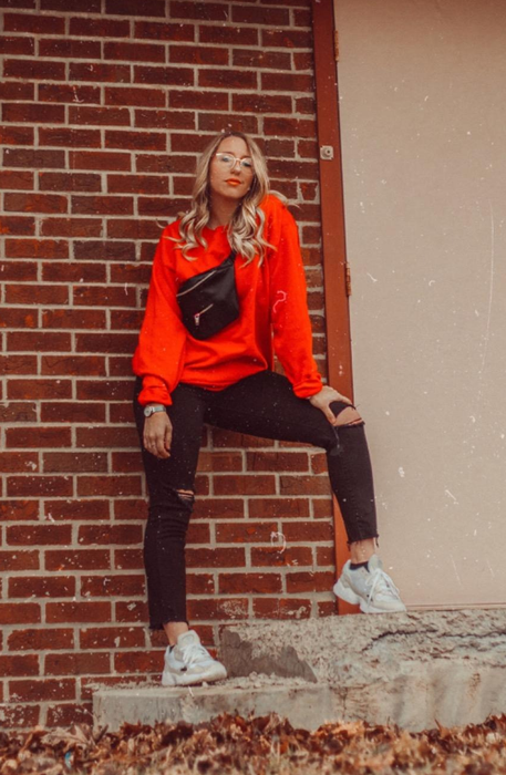 A reviewer posing against a brick wall, wearing a casual red sweater, black ripped jeans, white sneakers, and a black crossbody bag