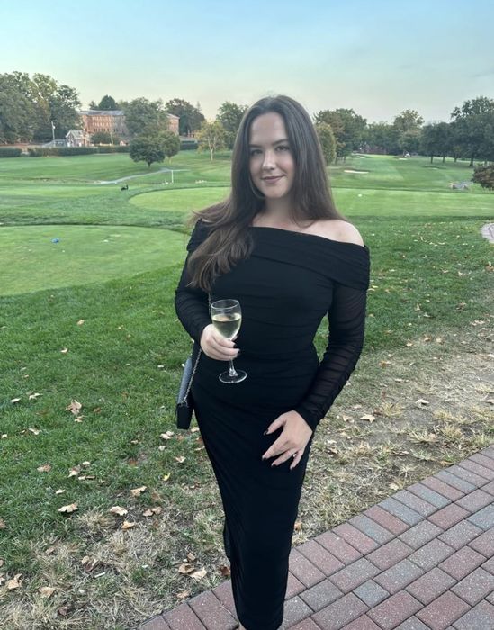 A reviewer wearing a sophisticated black off-the-shoulder dress poses outdoors, holding a wine glass with a lush green landscape in the background