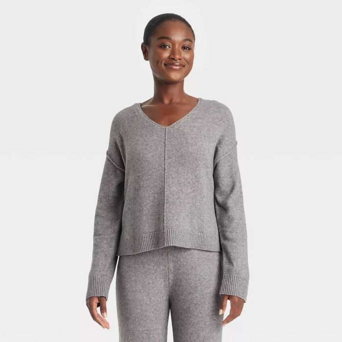 A person wearing a cozy, V-neck gray sweater and matching pants, smiling while standing against a simple backdrop.
