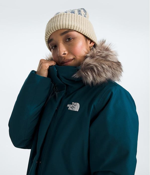Person wearing a teal fur-lined winter coat with a logo on the chest, paired with a knit beanie, featured in a shopping article