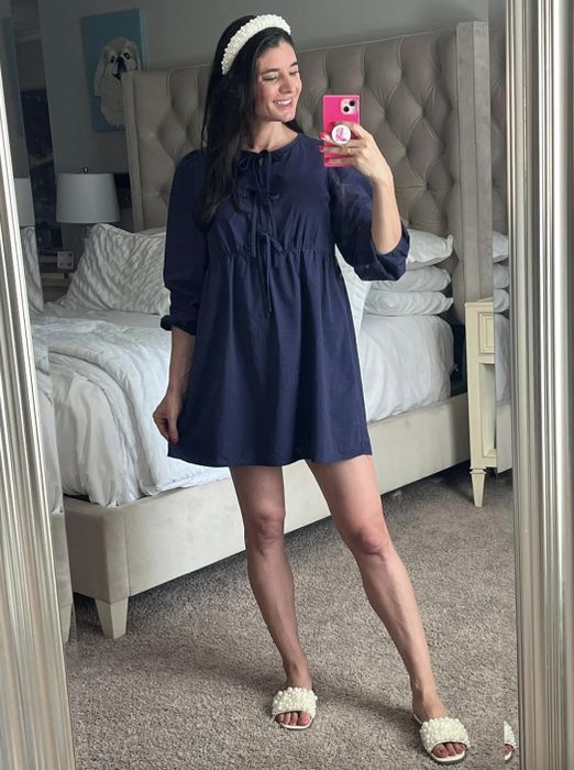 A reviewer captures a mirror selfie in her bedroom, dressed in a blue dress, a white headband, and pearl-detailed slippers. Her name is not provided