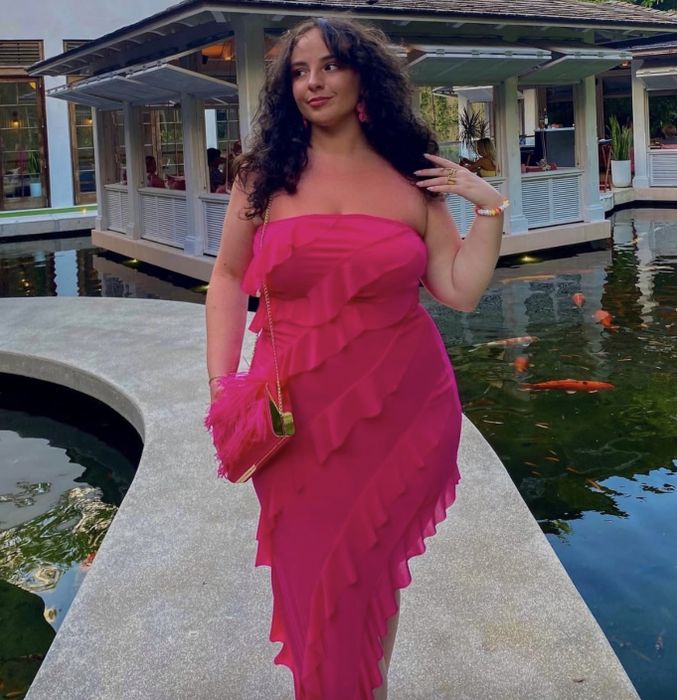 A woman in a ruffled strapless dress, carrying a pink handbag, stands gracefully by a pond filled with koi fish