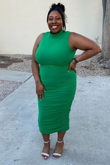 A reviewer smiles outdoors, dressed in a fitted sleeveless green dress paired with open-toe heels