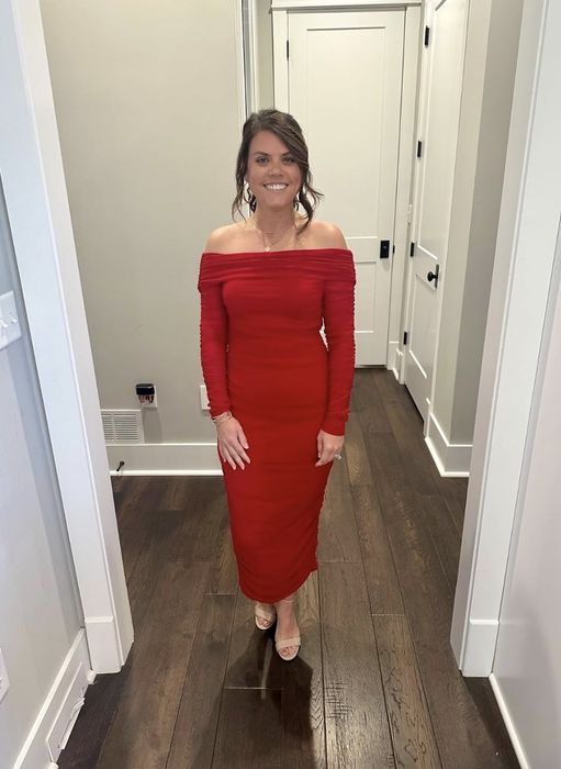 An individual in a stylish off-the-shoulder, long-sleeve dress stands in a hallway, smiling warmly at the camera