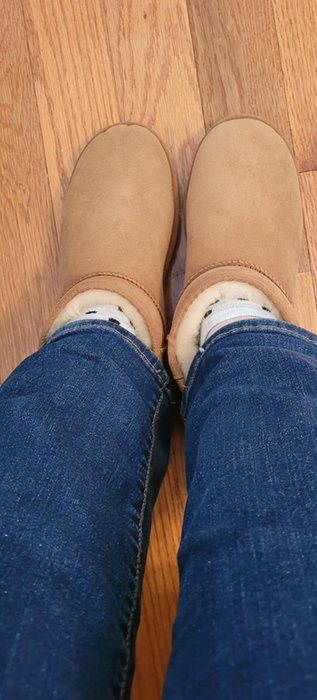 A person dressed in blue jeans and beige slippers stands on a wooden floor. The slippers look plush and perfect for indoor comfort