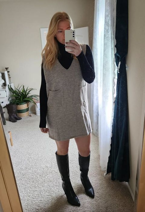 A person poses indoors, snapping a mirror selfie in a black turtleneck, a gray sleeveless dress, and knee-high black boots.