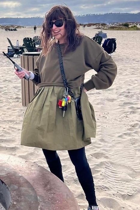 A person smiles on a beach, wearing an olive green babydoll dress, leggings, and sneakers with colorful laces, holding a lighter.