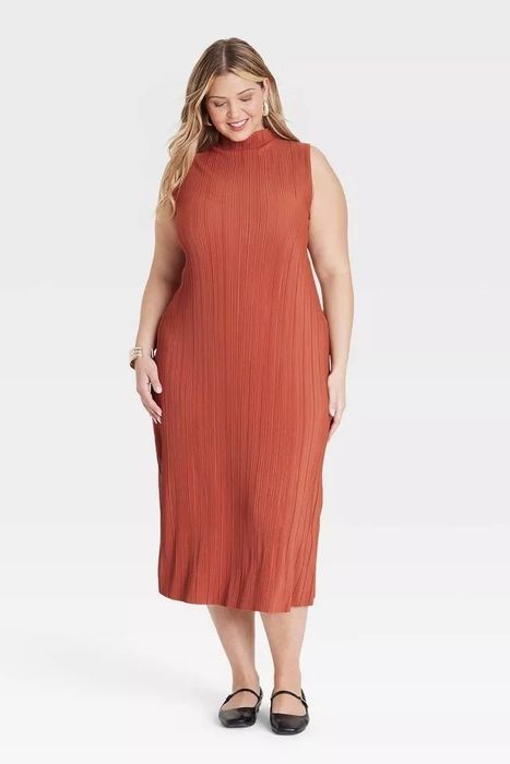 A model dressed in a sleeveless, ankle-length dress with a high neckline, smiling while looking down, accessorized with bracelets and black Mary Jane flats