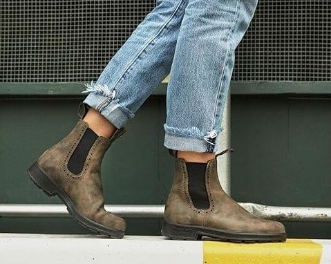 A person dressed in distressed jeans and brown ankle boots, standing confidently on a concrete barrier.