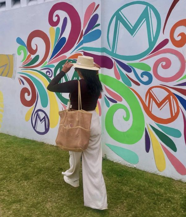 A person in white pants and a hat poses in front of a floral-patterned mural, holding a woven bag