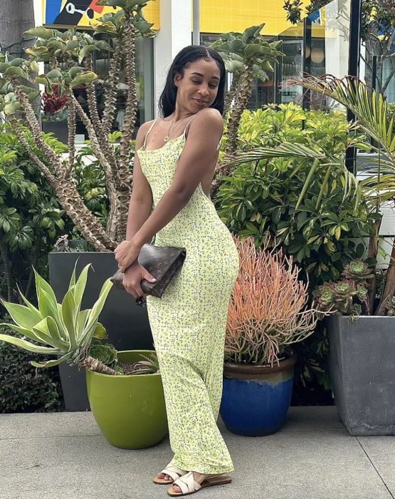 A woman in a fitted floral dress poses outdoors surrounded by lush greenery, smiling and holding a clutch
