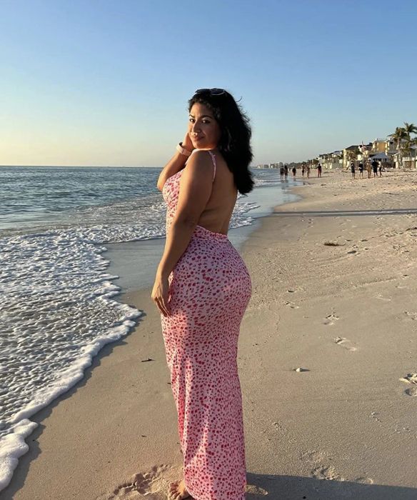 A woman in a sundress stands on a beach at sunset, waves gently touching the shore. She glances over her shoulder, her hand lightly touching her face