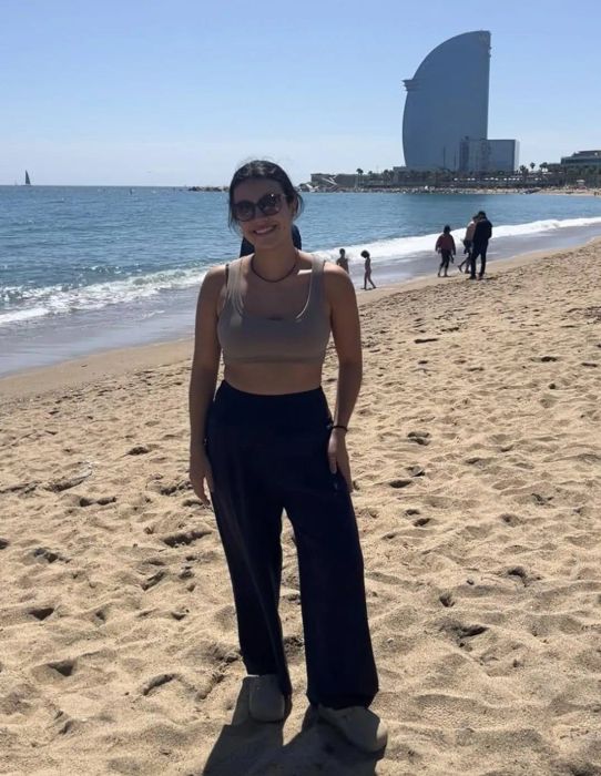 A reviewer wearing a tank top, sunglasses, and wide-leg pants stands on a beach with a modern structure in the background