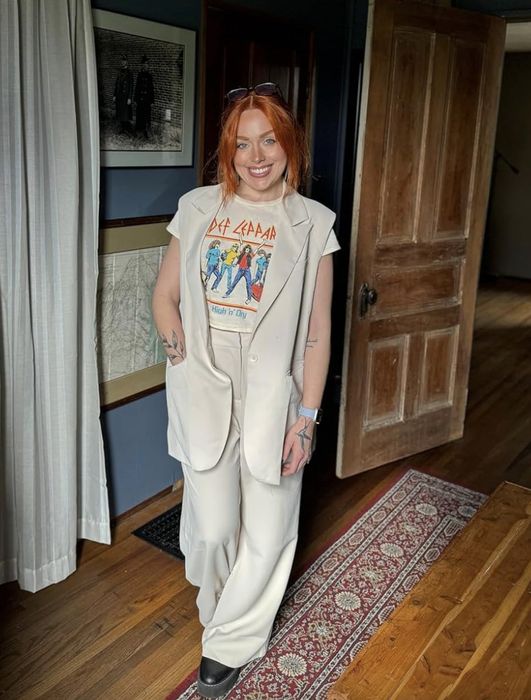 A reviewer with long red hair, wearing a Def Leppard t-shirt, a white vest, and wide-leg pants, stands in a room with hardwood floors and framed artwork.