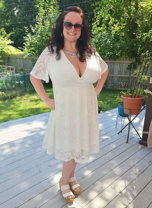 A person wearing a white lace dress, sunglasses, and a necklace, standing on a deck.