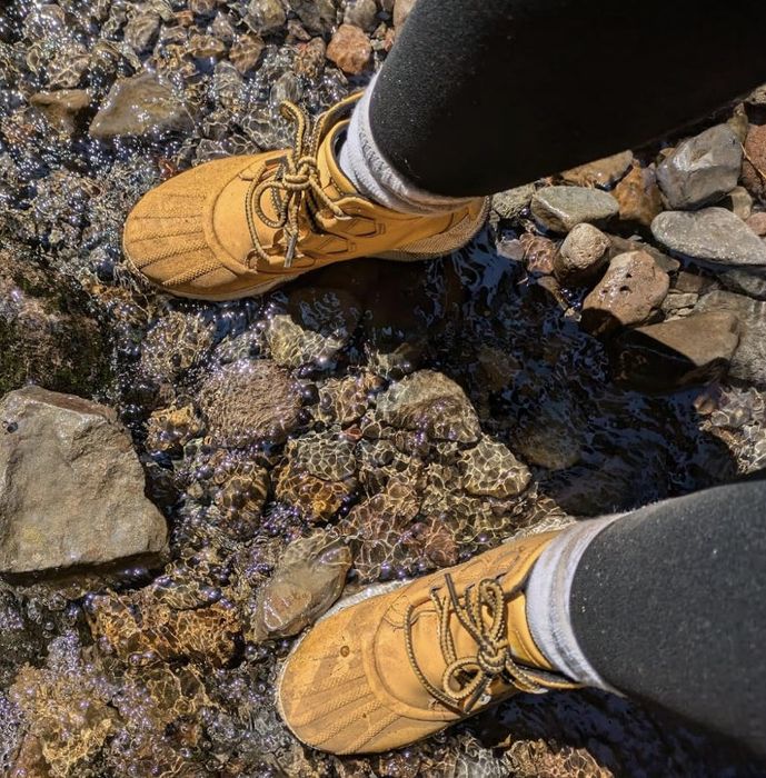 Tan moccasins worn by a person, positioned among rocks and shallow water, suggesting a nature walk or hiking environment.