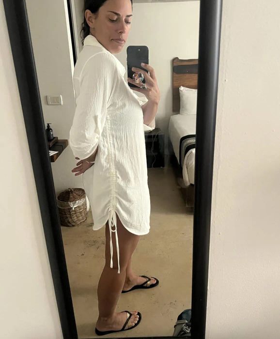 A person takes a mirror selfie wearing a casual, lightweight dress and flip-flops, standing in a room with a neatly made bed behind them
