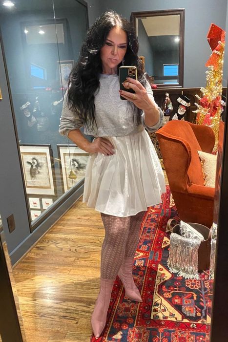 A reviewer takes a mirror selfie in a stylish gallery office, wearing a gray babydoll dress paired with pink boots.