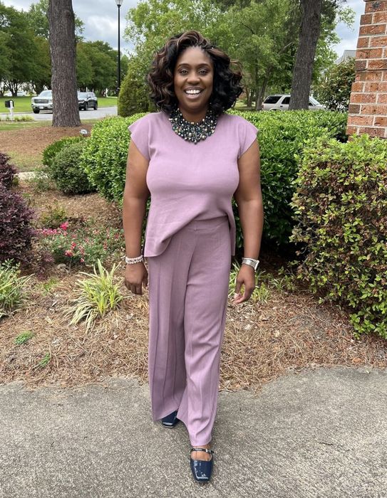 A reviewer stands outdoors, smiling, wearing a sleeveless, asymmetrical top with matching wide-leg pants and a statement necklace. The background features trees and bushes.