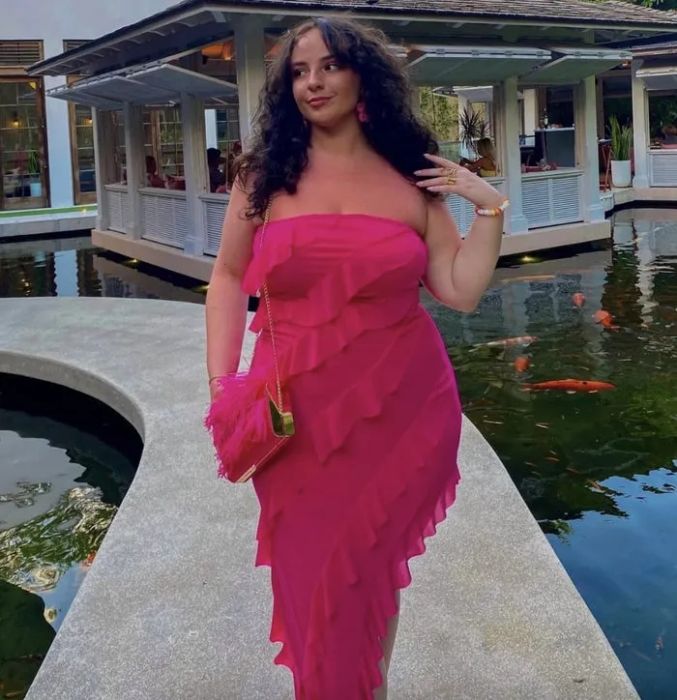 A woman wearing a strapless ruffled dress, carrying a pink handbag, stands by a koi fish pond