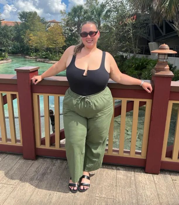 A person smiles outdoors, posing by a wooden railing. She’s wearing a sleeveless black top, green wide-leg pants, sunglasses, and sandals