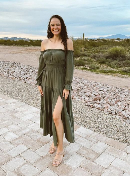 A reviewer strikes a pose outdoors in a long-sleeved, off-shoulder dress with a daring thigh-high slit, standing on a paved path with a desert backdrop.