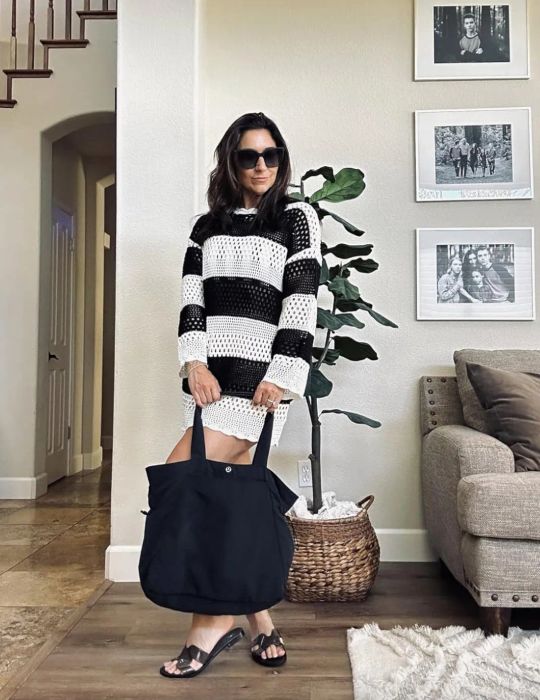 A reviewer in a black and white striped knit dress, sunglasses, and black sandals stands indoors holding a large black bag