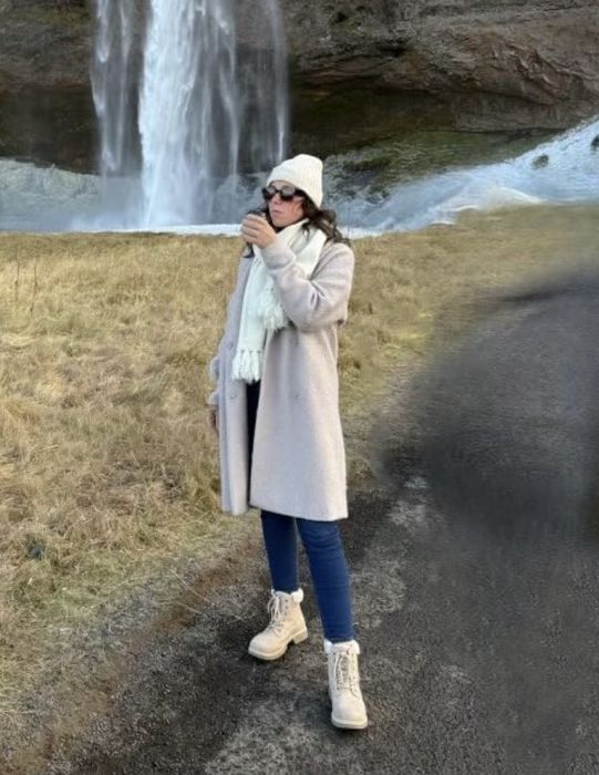 A person dressed in a coat and scarf stands by a waterfall, sipping from a cup. The landscape features grassy areas and rocky terrain in the background.