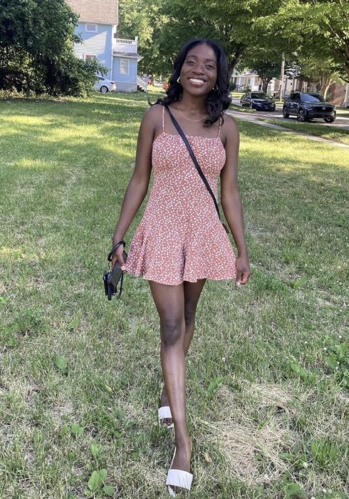 A reviewer smiles while strolling across a grassy lawn, donning a sleeveless floral dress, with a camera and crossbody bag in hand. Trees and houses can be seen in the background.